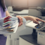 Bussinesswoman using copier machine to copy heap of paperwork in office.